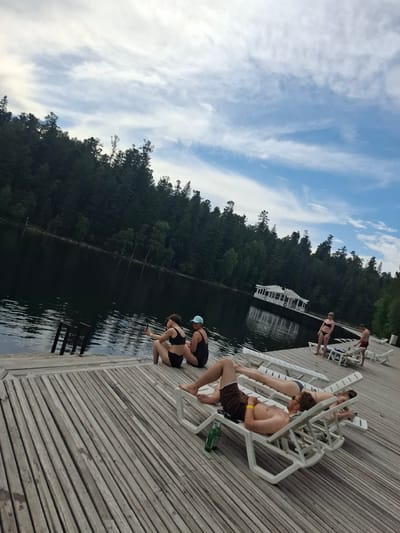 The image depicts a lakeside scene in Vydrino, Russia. A wooden dock extends into dark water, bordered by a dense forest. Several people are present. In the foreground, two individuals recline on lounge chairs on the dock. One male, wearing brown swim trunks, is lying on his back, while a female, in a bikini, lies next to him. Further down the dock, a woman in a black bikini and a blue hat sits with another woman in a black bikini. Two more individuals, a woman in a bikini and a man in swim trunks, stand near lounge chairs in the mid-ground. In the background, a white gazebo is situated on the water. The sky is partly cloudy with a blue and white color palette.
