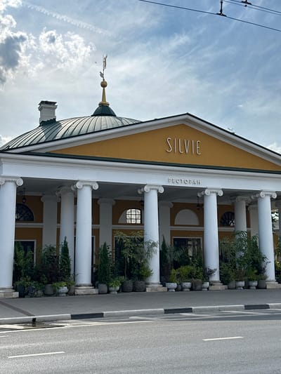 The image shows the facade of a restaurant called "SILVIE" in Golovinskoe, Russia. The building features a distinctive green domed roof with a golden weather vane topped by a figure. A white brick chimney is visible on the left side of the roof. The main entrance is adorned with a yellow facade, white columns with Corinthian capitals, and a prominent sign that reads "SILVIE" in large, metallic letters, with "РЕСТОРАН" (Restaurant) written below it in smaller letters. The building is surrounded by potted plants, which are arranged along the colonnade. A street with a white line marking the lane is visible in the foreground, with a sidewalk and curb separating it from the building. The sky above is partly cloudy with some wispy clouds and contrails. There are no people visible in the image.