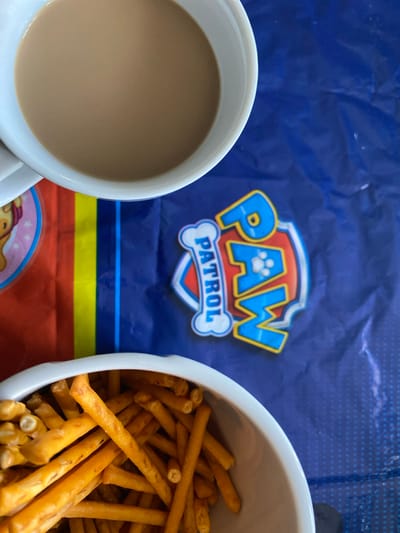 The image displays a close-up view of a table setting, likely at a child's birthday party, given the presence of Paw Patrol branding. On the left side, a white mug filled with a light brown beverage, possibly tea or coffee, is visible. Adjacent to it, a white bowl is overflowing with pretzel sticks. The surface beneath the objects is a blue, crinkled plastic mat featuring a prominent Paw Patrol logo, which includes the words "PAW" and "PATROL" in bold lettering. The mat also has sections with red, yellow, and blue stripes. The overall scene suggests a casual, indoor setting, potentially a home environment. The image does not provide any information about the city of Osek, Czechia, or any individuals present.