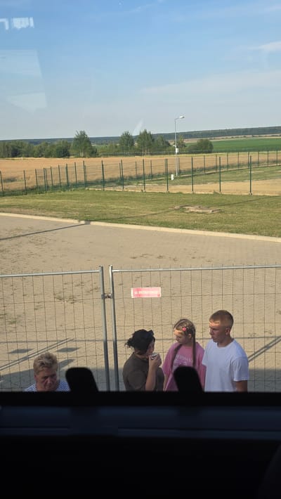 In the city of Dołhobyczów, Poland, four individuals are visible behind a temporary metal fence. On the far left, an elderly woman with blonde hair is looking down. Next to her, a woman with dark hair tied back is drinking from a mug. A young girl with braided hair, wearing a pink shirt with the text "ROISSANT," is looking towards the woman with the mug. To the right of the girl, a young man with short blonde hair is wearing a white t-shirt and looking to the side. The fence has a red sign attached to it that reads "BETAFENCE". In the background, a field of dry grass is bordered by a fence and trees, with a street lamp visible on the right. The sky is clear and blue. The image appears to have been taken from inside a vehicle, as indicated by the dark interior at the bottom of the frame.