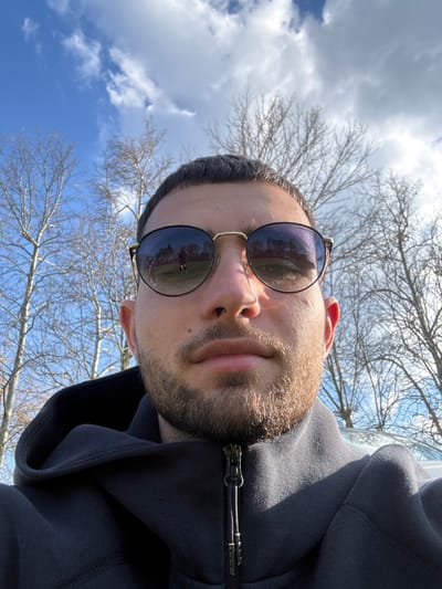 A man is the subject of the photograph. He is wearing sunglasses, and a black hooded sweatshirt. The background features a blue sky with white clouds and bare tree branches. The reflection in his sunglasses contains people standing on a field of green. The exact location in the city of Dushevo, Bulgaria is indeterminable from the image alone.