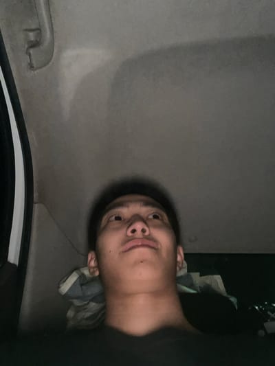 The image shows a low-angle, dimly lit shot of a person's face looking up from inside a vehicle. The person appears to be young, with dark hair and a light complexion. They are wearing a dark shirt. The interior of the vehicle is visible, with a grey headliner and a part of the door frame on the left. The background is dark and out of focus, with some indistinct shapes that could be luggage or other items. The lighting suggests it is nighttime or very early morning. The specific location in Aytey, Kazakhstan, cannot be determined from the image alone.