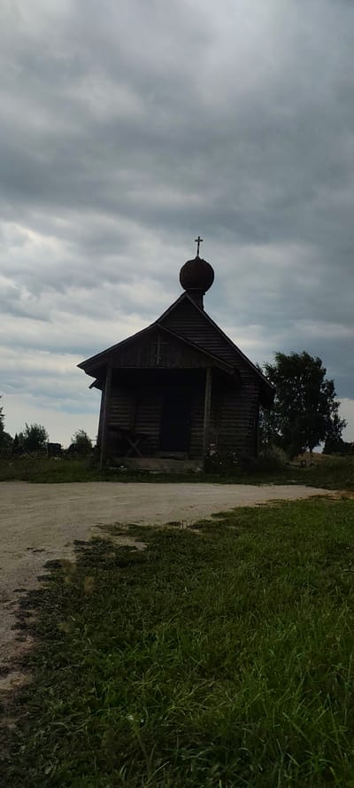 A small, dark wooden chapel with a rounded dome and a cross on top stands in the foreground under a cloudy sky. The chapel has a sloped roof and an open entrance. A dirt path leads to the chapel, with grass and trees in the background. The overall scene is muted and somewhat somber due to the overcast weather. This chapel is located in the historic fortress of Izborsk, in the city of Novy Izborsk, Russia.