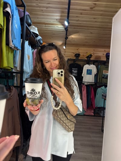 A woman is taking a selfie in a clothing store. She is wearing a white t-shirt and black pants. She is holding a coffee cup in her left hand and her phone in her right hand. The coffee cup has a picture of a tree and the word "КОФЕ" (coffee in Russian) on it. She is also wearing a pearl necklace and a Gucci bag. The store is filled with clothes, hats, and other merchandise. The walls and ceiling are made of wood. The floor is made of laminate. The scene appears to be in T/B Seminskii Pereval, Russia.