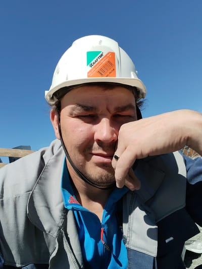 This is a selfie of a man wearing a white hard hat and work clothes, standing outdoors against a bright blue sky. The man has a beard and is resting his hand on his chin, with his eyes partially closed, suggesting he may be tired or squinting in the sun. He is wearing a grey work jacket over a blue shirt, possibly with an Adidas logo visible on the collar. The hard hat has a sticker with the brand name "KRAFTER" and text in Russian and Kazakh, indicating it is a protective helmet. The background shows a clear, sunny sky and hints of construction or outdoor work with wooden beams and some buildings in the distance. The overall impression is of a construction worker taking a break on a bright, sunny day.