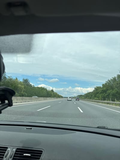 The image shows a view from inside a car traveling on a highway, with the perspective slightly angled upwards. The highway is divided by white lines, and two vehicles can be seen ahead. Trees line the sides of the highway, with a blue sky and scattered clouds visible above. The car's interior is partially visible on the left and right sides of the frame. It is not possible to verify the location as Fischamend, Austria from the image.