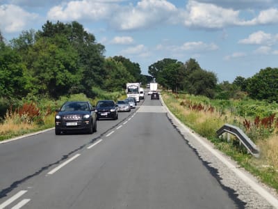 The image depicts a traffic jam on a two-lane asphalt road, likely in or near Zgalevo, Bulgaria, as indicated by the car license plates. The scene is set during the daytime with a partly cloudy sky. A line of vehicles is visible, including a black Jeep Compass in the foreground, followed by several other cars and two white trucks further down the road. The road is bordered by a grassy verge and trees on both sides, with some dry vegetation present. A guardrail is visible on the right side of the road. The cars appear to be stopped or moving very slowly, suggesting a significant traffic delay.