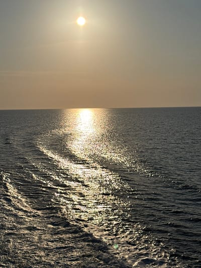 The image displays a wide view of an open body of water under a bright sky, characteristic of a marine environment. The sun, a radiant, star-like point of light, is positioned in the upper portion of the frame, creating a strong, shimmering reflection path that extends across the water's surface from the horizon towards the bottom center. The sky above is a pale yellow-orange hue, suggesting either sunrise or sunset. The horizon line is flat and clearly defined, dividing the sky from the water. The water itself is dark blue-grey, disturbed by a distinct wake originating from the lower-left foreground and extending diagonally towards the mid-right. This wake consists of undulating waves and foam, indicative of a vessel's recent passage. No specific geographical features or individuals are visible. If this image were captured from Kato Karioutes, Greece, it would depict the coastal waters adjacent to that city.
