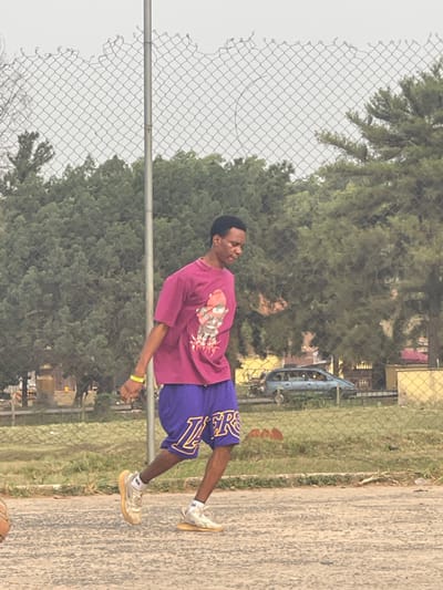 An outdoor scene in Umunekwu, Nigeria, features a young Black male standing on a paved court. He is positioned in a mid-stride or dynamic pose, leaning slightly forward and to his right, with his left foot elevated. He wears a magenta short-sleeved t-shirt with a multi-colored graphic, purple basketball shorts displaying 'LAKERS' in yellow text, and light-toned athletic shoes. A yellow wristband is on his left wrist. A basketball is partially visible on the ground in the lower-left foreground. A grey wire mesh fence, with an uneven top edge and a vertical metal post, spans the midground. Beyond the fence, a dense collection of green trees fills the background. In the further distance, a light blue or grey vehicle and faint building structures are visible under an overcast sky.