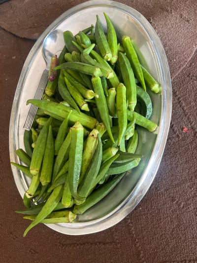 A close-up, overhead shot shows a metal oval platter filled with fresh, green okra pods. The okra is piled high, with some pods overlapping and others lying at various angles. A silver spoon rests on top of the okra, near the left edge of the platter. The platter itself is placed on a textured brown surface, possibly a tablecloth or a rug. There are no people visible in the image. The lighting appears natural, casting soft shadows on the okra. The setting is simple and domestic. The location in Kabul, Afghanistan is not discernible from the visual information provided in the image.
