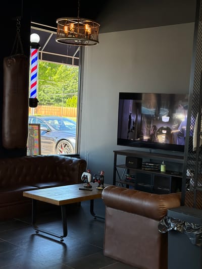 The image depicts the interior of a modern barber shop with an open garage door. In the foreground, a traditional red, white, and blue barber pole is illuminated. To the left, a decorative industrial-style light fixture with exposed bulbs hangs from the ceiling. A dark brown leather sofa occupies the lower portion of the image, with a punching bag suspended by chains behind it.
In the mid-ground, through the open garage door, a silver car is visible parked on a tree-lined street. The interior of the shop features a dark gray wall and black tiled flooring. A flat-screen television mounted on the wall displays a scene from an airplane cabin. A dark brown leather armchair is situated to the right of the television. A small wooden table with black metal legs sits in front of the armchair. On the table, there are a few items, including what appears to be a white game controller. A bookshelf unit behind the television holds several books and decorative items. The overall atmosphere suggests a contemporary and stylish barbershop.
The time of day appears to be daytime, with natural light streaming in through the open garage door. The weather seems pleasant, indicated by the lush green trees visible outside. No specific emotions are discernible from the image. There is text visible on the barber pole, which reads "208" and "BARBER SHOP" in reverse.