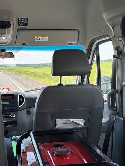 The image depicts the interior of a vehicle, likely a van or bus, with a focus on the passenger seating area and dashboard. In the foreground, a red portable generator is visible, secured by black metal bars. To the left of the generator, a plastic water bottle sits on a green object, possibly a mat or bag.
Above the dashboard, a sun visor is partially visible, with an "AIRBAG" warning sticker and other symbols. On the ceiling of the vehicle, above the sun visor, a panel with multiple icons is affixed, indicating seating arrangements possibly related to passenger capacity or accessibility features, including icons for wheelchairs and seated individuals.
The driver's side of the dashboard is visible, showing the steering wheel and various controls. The view through the windshield reveals a rural landscape with a road, green fields, and trees under a cloudy sky. The interior of the vehicle appears to be clean and functional. The image does not contain any individuals. The image context suggests the vehicle may be in or traveling through the Мастоцкі сельскі Савет region of Belarus, based on the user prompt.
