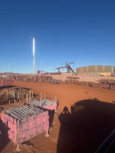 The image captures a vast, open outdoor industrial storage yard under a clear blue sky. The ground is composed of reddish-brown dirt, characteristic of the Australian outback. In the foreground, several stacks of metal components are organized and prepared for transport or use. These components appear to be part of a construction or scaffolding system, some wrapped in pink protective material.
In the mid-ground, rows of metal poles and frames are neatly stacked, indicating a well-managed inventory. Beyond this, industrial structures are visible, including large cranes and a long, green and tan warehouse-style building. A prominent tall pole with bright lights stands in the distance, reflecting intensely due to the sunlight. The scene suggests an active industrial site, likely involved in mining or construction operations, potentially in or near Newman, Australia, given the characteristic red earth and industrial infrastructure. A shadow of a person and possibly a vehicle is cast across the foreground, indicating the presence of activity at the time the photograph was taken.