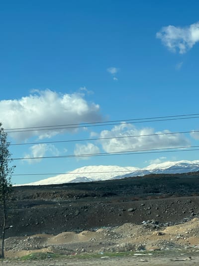 The image displays a panoramic outdoor scene during daylight, located in Al-Kiswa, Syria. The upper portion of the frame features a clear blue sky interspersed with scattered white cumulus clouds. Four parallel, dark-colored power lines horizontally traverse the middle of the image, partially obstructing the view of the background.

In the distance, a range of mountains is visible, with their upper slopes and peaks covered in white snow. Below the snowline, the mountains and hills transition into dark, rugged terrain, appearing barren and rocky.

The midground consists of an expanse of dark, uneven ground with scattered rocks and debris. The foreground features lighter, sandy-colored earth with occasional patches of sparse green vegetation. Several small, irregular mounds of sand or soil are present in the foreground. Visible litter, including plastic bags and miscellaneous debris, is scattered across the foreground terrain. A slender, partially obscured tree trunk with some branches is present on the far left edge of the foreground. No human figures or discernible activities are present.