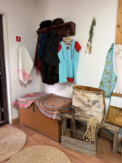 This is a medium shot, eye-level, interior photograph taken in a room in Ust'-Kabyrza, Russia, likely part of a museum or historical exhibit. The scene depicts a collection of rustic, traditional items displayed against a white wall and wooden floor.

On the wall, a dark wooden shelf holds two dark, fur hats. Below the shelf, a wooden peg rack has several items of clothing hanging: a red and black patterned scarf, a dark blue scarf, a black vest over a dark brown garment, and a light blue dress with red trim and embroidered details. To the left of the rack, a white linen cloth with pink trim is draped. To the right, a dried bunch of herbs hangs on the wall. Further to the right, a light blue apron with a strawberry pattern is hung.

In the foreground, a large, wooden chest sits on the floor, partially covered by two round, woven mats with concentric circular patterns in various colors, including orange, pink, and gray. To the right of the chest, a rustic wooden bench supports a large, upright birch bark basket. A weathered wooden toolbox sits on the floor in front of the bench, containing a rusty metal tool with a wooden handle and a coiled rope. A woven straw basket is visible on the right side of the bench. Two round, woven straw mats are on the floor in the lower left corner of the image. A red square sign with a white fire extinguisher symbol is mounted on the wall to the left of the clothing rack. The overall impression is one of a preserved domestic or workshop setting.