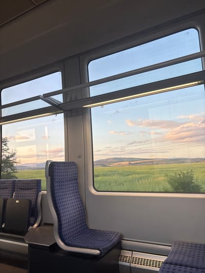 The image depicts the interior of a passenger train car, looking out through its windows at a rural landscape during what appears to be late afternoon or early evening. The scene is described as taking place in Tsar Asen, Bulgaria.
No individuals are present in the train car. The interior features a light gray ceiling, with a textured, possibly ribbed, section on the upper left and a smooth surface dominating the rest. Linear artificial light fixtures are active along the ceiling, casting subtle reflections onto the windows.
Multiple large windows frame the exterior view. On the left side, two vertically arranged windows show a partial, slightly blurred view of a green tree close to the train, with distant rolling hills and a soft, cloudy sky illuminated by warm, golden light. Reflections of the train's interior, including the ceiling and light fixtures, are prominent in these windows.
The primary exterior view is through a large, rectangular window on the right, which dominates the frame. This window overlooks an expansive, vibrant green agricultural field in the immediate foreground and midground. Beyond the field, a series of softer, undulating hills recede into the distance under a pale blue sky streaked with diffuse pink, orange, and white clouds, characteristic of sunset. Several parallel overhead power lines or communication lines are visible crossing horizontally across the sky in the upper portion of the exterior view. A small, dark bush or clump of trees is visible in the lower right corner of the field.
The train's interior is furnished with blue passenger seats featuring a small, repeating grid pattern. These seats have light gray, curvilinear frames. A prominent empty seat in the mid-right foreground faces away from the camera, with a small, dark gray fold-down table extended from its back. To the far left, the backs of two other blue seats are partially visible, one featuring a black tray table with the number '9' printed on it. A vertical structural pillar between the right-side windows displays a white sticker with the number '55' in black text. The floor is dark. A metallic grille, likely for heating or ventilation, is situated at the base of the wall below the rightmost window.
The overall illumination combines the warm, fading natural light from outside with the cooler, artificial lighting inside the train, creating a serene and transitional atmosphere.