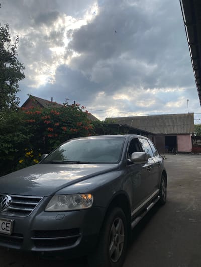The image captures a gray Volkswagen Touareg parked on a paved surface in front of a building. The building appears to be a workshop or garage with a corrugated roof. Behind the car and to the left, there is a red-roofed structure partially obscured by a dense bush with orange flowers. Yellow flowers are visible below the orange-flowered bush. The sky is overcast with dramatic clouds. A single small, dark object is visible in the sky to the upper right of the frame, possibly a bird or insect. The overall scene suggests a rural or semi-rural setting.