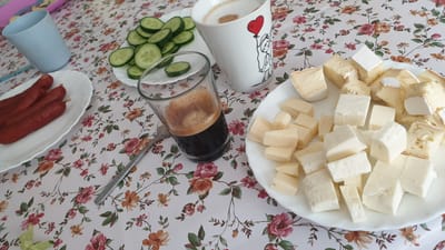 A close-up, eye-level shot shows a breakfast spread on a floral-patterned tablecloth. In the foreground, a white plate holds an assortment of cubed cheeses, some with visible rind. To the left, a small glass contains dark liquid, likely coffee, with a fork resting beside it. Further left, a white plate features several elongated, reddish-brown sausages. Behind the sausages, a light blue plastic cup stands. A white mug with a simple drawing of a person holding a red heart is visible in the upper center. Adjacent to the mug, a small white plate is piled with thin slices of cucumber. The scene is set indoors, with the lighting suggesting it could be morning or daytime. No people are visible in the image.