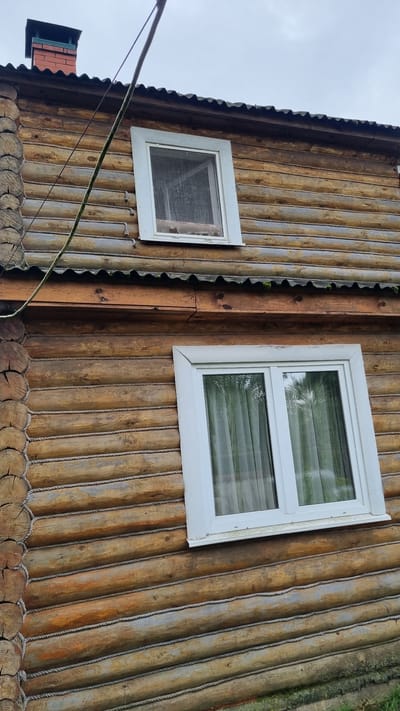 The image shows the exterior wall of a log cabin in Солнечная Поляна-3 Снт, Russia. The wall is constructed from horizontal logs, with rope detailing filling the gaps between them. There are two white-framed windows visible on the wall. The upper window is smaller and appears to have a screen. The lower window is larger and is covered by light-colored curtains. Above the windows, a corrugated roofline is visible, and a brick chimney with a black cap protrudes from the roof. A power line runs diagonally across the upper portion of the image. The overall impression is of a rustic, rural dwelling.