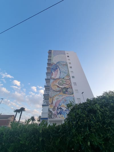 The image captures a portion of a building in La Pobla de Farnals, Spain, notable for its large, colorful mosaic mural adorning the side. The building is white with a series of balconies along the side where the mural is located. The mural itself features swirling patterns in shades of blue, pink, yellow, and white, made from individual tiles. The sky is a clear blue, and partially obscures the top of the building. A power line runs across the left side of the image. The right side of the frame is dominated by a large, green tree, partially obscuring other buildings and palm trees behind it. The angle of the shot is from below, looking upwards at the building. Some faint clouds can be seen near the buildings in the background.