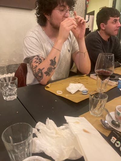 The image depicts an indoor scene, likely a casual restaurant or bar in Sondrio, Italy, featuring two young men seated at a dark table. A brown paper runner or placemat is spread across a portion of the table.
In the foreground, a man with dark, curly hair, glasses, and a beard is actively drinking from a clear glass, which he holds to his mouth. He is wearing a light grey short-sleeved t-shirt, showcasing several tattoos on his left arm, including a prominent rose, an anchor, and other designs. A thin bracelet is visible on his right wrist.
Behind him, another man with short, dark hair and an earring is visible, wearing a dark long-sleeved top. He is looking upwards and to the right, away from the camera.
The table is set with various items suggesting a meal or social gathering. A glass carafe filled with red wine stands next to an empty clear drinking glass. Several crumpled white paper napkins, some stained, are scattered on the table and the paper runner. A small blue bottle cap and a white plastic ring are also present. Further right, a coffee cup resting on a saucer with spoons indicates a finished beverage. A pack of cigarettes with "ULTRA" visible on its blue and white packaging is placed on the table. Another empty clear drinking glass is situated in the bottom right corner of the frame. The scene conveys a relaxed, casual atmosphere, characteristic of an evening outing.