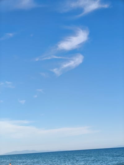 This is a vertical, eye-level shot of a serene seascape at Castiglione della Pescaia, Italy. The image is dominated by a vast expanse of clear blue sky, adorned with wispy white clouds that form a shape resembling a stylized bird or a crescent moon. In the distance, the calm, blue sea stretches towards a hazy horizon, where faint, undulating hills are visible. The water is slightly rippled, catching the light and creating a gentle shimmer. A small portion of the scene at the bottom left reveals a hint of a beach with two people visible in the water, one wearing an orange float. The overall atmosphere is one of tranquility and natural beauty.