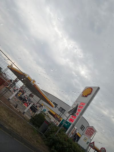 The image captures a Shell gas station located in Lohne, Germany, under a cloudy sky, with visible raindrops on the lens creating a blurred effect. The gas station features a prominent Shell logo and a price display board showing fuel prices: 224.9, 213.9, 19.9, 17.9, 223.9. Adjacent to the main structure, a smaller sign displays "JETZT AUFLADEN". The scene appears to be under construction or renovation, with fencing and construction materials present, including red and white striped barriers. The overall environment is an urban outdoor setting, typical of a service station area. No individuals are clearly visible in the frame.