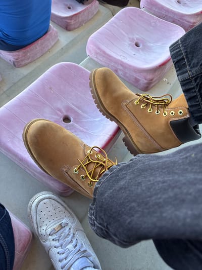The image presents a low-angle, eye-level shot of a person seated in a stadium setting. The individual's feet, clad in two different types of footwear, are prominently displayed. One foot wears a tan-colored Timberland-style work boot with yellow and burgundy laces, while the other sports a white Nike Air Force 1 sneaker. The person is wearing dark denim jeans.
The seating appears to be made of molded pink plastic, with several rows visible in the background. The setting suggests an outdoor stadium or arena.
While the exact location within Valpaços, Portugal cannot be determined solely from the image, the stadium seating suggests a sports facility or a similar public gathering space within the city. No other individuals or specific landmarks are visible in the frame to pinpoint the location further.
