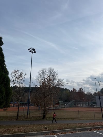 A landscape shows a partly cloudy sky above a park. There are trees with bare branches, and a person is walking along a path next to a baseball field. Tall light poles stand near the trees. The edge of a paved road is visible on the right. The overall scene evokes a sense of autumn or winter with the subdued colors and bare trees.