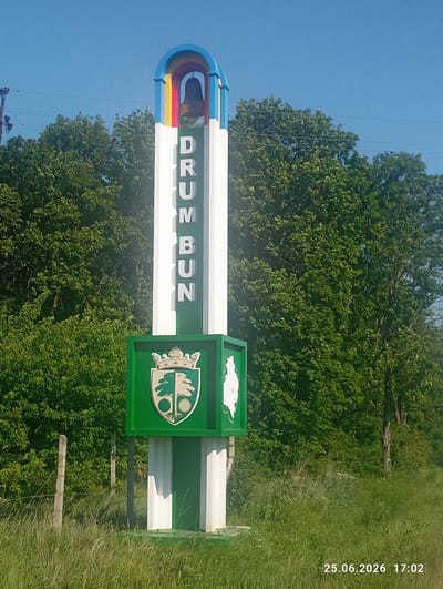 The image displays a roadside monument in Cojușna, Moldova. The monument is tall and white with green and white stripes at the base. A decorative arched top in blue, yellow, and red frames a bell. Vertical green letters on the white surface spell out "DRUM BUN," which translates to "Good Road." Below the text, a hexagonal green section features a coat of arms on its front face, depicting a crown above a shield split in half vertically, with a green tree with circular fruit on the left and a similar tree on the right. To the right of this is another green hexagonal section with a white silhouette of a tree. The monument is surrounded by lush green trees and vegetation, with a clear blue sky above. In the foreground, tall grass and a barbed-wire fence are visible. The lower right corner of the image contains a timestamp: "25.06.2026 17:02".