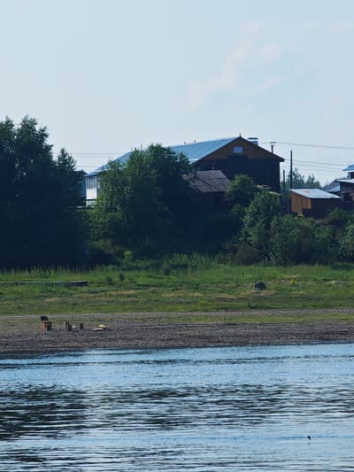 The image depicts a waterfront scene in Verkhnepashino, Russia, on a clear day. In the foreground, a body of blue water with ripples covers the lower portion of the frame. Along the shore, a narrow strip of grey pebbles is visible, transitioning to green grass and foliage further inland. Several wooden structures, appearing to be benches or small tables, are scattered on the pebble area.
Behind the waterfront, a slope is covered with lush green vegetation and trees. Dominating the middle ground are several buildings with pitched roofs. One large, dark-colored building with a yellow-brown upper section and a blue roof is prominent, featuring a chimney and a small window. To its left, a smaller building with a corrugated roof is visible. To the right, a smaller, wooden outbuilding with a corrugated metal roof stands. More trees and vegetation surround these structures, with power lines visible in the background. The sky above is pale blue with a few wispy clouds. There are no visible people in the scene.