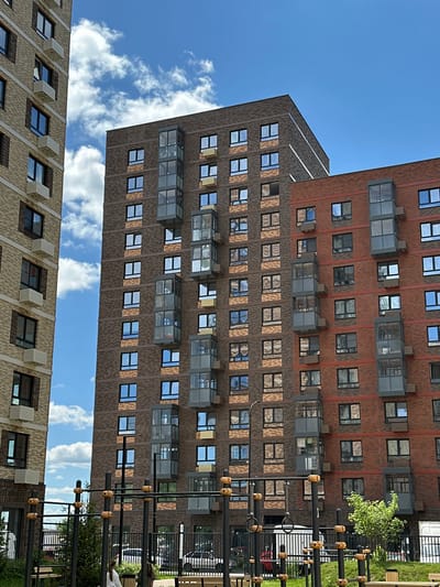 In Znamya Oktyabrya, Russia, this image captures a segment of a residential complex with a clear blue sky dotted with white clouds. Three multi-story buildings are visible, two of which are tall apartment buildings. The building on the far left is clad in light-colored brick, featuring regularly spaced windows and what appear to be air conditioning units mounted below some of them. The central building is taller and clad in darker brown brick, with windows arranged in a grid pattern. It also has several enclosed balconies with glass fronts and gray frames, stacked vertically. The building to the right is constructed with red brick, similar in height to the central building, and also features enclosed balconies. In the foreground, a black metal outdoor fitness or playground structure with wooden grips is prominent. A black metal fence runs horizontally across the lower portion of the image, separating the play area from a street or pathway where parked vehicles are visible, including a white van and a light-colored car. Two young females are in the foreground, one facing away from the camera and the other partially visible. Green grass and a small tree are present in the immediate foreground.