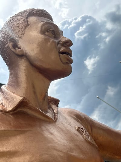 A low-angle close-up shot focuses on the bronze bust of a man, looking upwards towards a cloudy sky. The sculpture appears to be part of an outdoor installation, possibly a monument or artwork, given the reflections of overhead lights and cables visible against the sky. The statue's surface is a warm, metallic bronze, showing details of the man's facial features, including his lips, nose, and the textured appearance of his hair. The cloudy sky suggests it is daytime, though the exact time is indeterminate. There is no visible text within the frame. The scene is set outdoors, with the sky and clouds forming the backdrop for the sculpted figure. The upward gaze of the statue could convey a sense of hope, aspiration, or contemplation. The image is oriented such that the sky occupies the top portion, and the sculpture dominates the foreground.