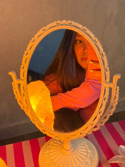 A young woman is taking a selfie in a decorative vanity mirror. She is wearing a pink long-sleeved shirt and has her hair down. Her left arm is visible, holding a bright orange cell phone. A Himalayan salt lamp, glowing orange, is positioned to the left of the mirror, also reflected within it. The mirror itself is framed in an ornate, cream-colored stand with intricate carvings. The background wall appears textured and grey. In the foreground, a striped pink and white mat is visible. The image is lit by the warm glow of the salt lamp and possibly indoor lighting, casting an amber hue over the scene. The specific location in the city of Prikubanskiy, Russia, cannot be determined from the visual information provided in the image.