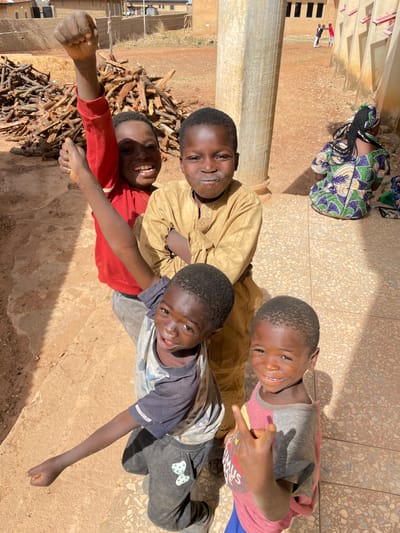 Four boys are lying down outdoors on a textured, light brown surface, possibly a patio or tiled area. The boy on the left is wearing a red shirt and has his fist clenched in the air. Next to him is a pile of wood. The boy in the middle is wearing a tan shirt, and the boy to his right is wearing a grey and white shirt with dark pants. The boy on the top right is wearing a grey and pink shirt and is holding up a hand gesture. Behind them is a light brown wall or building, with a cylindrical column visible. In the background, there is a glimpse of a building with dark windows and a person kneeling beside a colorful bag. The scene appears to be in Rahwol Chom, Nigeria.