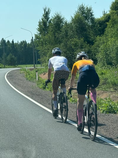 Two individuals are cycling on a paved road in a rural, tree-lined area, likely on the outskirts of Ермилово, Russia. The cyclists, viewed from behind, are wearing helmets and cycling attire. The person in front is wearing a white t-shirt and leopard-print shorts, while the person behind is wearing a colorful yellow and orange top and black cycling shorts, complemented by pink compression socks. Both are riding road bikes with black frames and spoked wheels. The road curves to the left, bordered by a gravel shoulder and lush green vegetation. In the background, a dense forest can be seen under a clear blue sky. Streetlights are visible along the roadside.