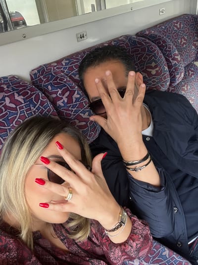 The image depicts two individuals, a female and a male, seated side-by-side inside a train cabin, consistent with a journey through or to Sidi Yahya El Gharb, Morocco. Both are wearing sunglasses and are actively holding their left hands up, fingers spread, to obscure portions of their faces, specifically their eyes, while being photographed.
The female, positioned on the left, has shoulder-length blonde hair with darker roots and manicured nails painted in a bright red hue. She is wearing a patterned top with shades of burgundy and beige. Her left hand displays a multi-band gold-colored ring on her ring finger, and her left wrist is adorned with multiple bracelets, including a wide, possibly metallic, piece and a thinner, silver-toned chain bracelet.
The male, situated to the right of the female, has short dark hair and is dressed in a dark blue collared jacket over a lighter-colored undershirt. His left wrist features three distinct bracelets: a black braided leather bracelet, a silver-toned metallic bracelet with a textured or coiled design, and another silver-toned bracelet accented with an anchor motif.
The train cabin environment includes upholstered seats covered in a repeating geometric pattern of blue, red, and beige. White paneling is visible above the seats, bearing black numerical identifiers '44' and '42', indicating seat numbers. A reflective surface in the upper left background shows a blurred exterior scene, including a portion of a red vehicle and indistinct vegetation, suggesting the train is in motion.