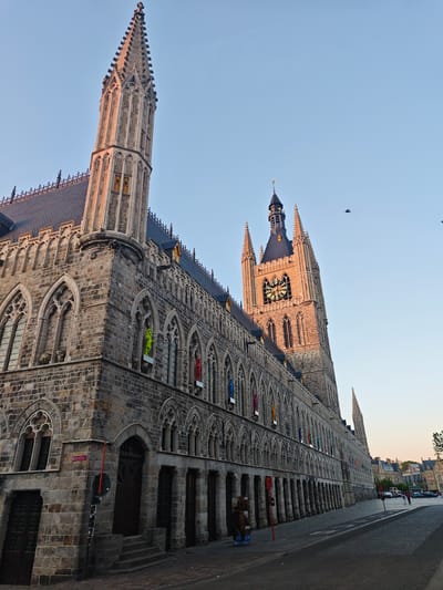 The image captures a grand, historic Gothic-style building, identified as the Cloth Hall (Lakenhalle) in Ypres, Belgium, seen from a low-angle perspective looking upwards. The building is constructed from light-colored stone blocks and features intricate architectural details. On the left, a tall, slender spire rises prominently, adorned with pinnacles and arched openings. On the right, a larger, more robust belfry (bell tower) stands with a visible clock face, also featuring ornate Gothic elements and crowned with a dark, pointed roof.
The main facade of the building stretches along a paved street, displaying numerous arched windows, many with delicate tracery. Along the ground floor, an arcade of stone columns creates a sheltered walkway. The roof is steeply pitched and covered with dark tiles. The warm, golden light illuminating parts of the building, especially the towers and upper sections, indicates either sunrise or sunset (golden hour). The sky above is clear and a soft, light blue.
Small birds are visible flying in the clear sky. Along the street, a few people can be seen walking in the distance, and several cars are parked further down. Notable details include colorful, human-like figures or banners hanging from some of the upper windows, with a bright green one and a red one clearly visible. Near the street, there are red poles and newspaper vending boxes. A red sign on the corner of the building prominently displays the text "Lakenhalle" and "Hall aux Draps," confirming its name and location. The scene conveys a sense of historical grandeur in a peaceful city setting.