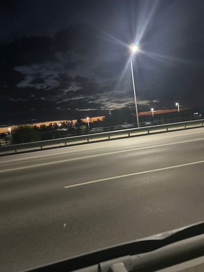 The image captures a nighttime scene from an elevated perspective, likely a bridge or overpass, looking across a multi-lane road. The road surface is asphalt with white lane markings visible. To the left, a metallic guardrail runs along the edge of the road. Beyond the guardrail, there is a strip of vegetation followed by a distant cityscape illuminated by numerous lights. The sky is dark, with scattered clouds and a prominent bright streetlamp in the upper right quadrant, casting rays of light. The horizon displays a band of orange light, possibly from a sunset or artificial lighting in the distance. The overall impression is one of a highway environment at dusk or night in an urban or semi-urban area, consistent with the location of Заречный ауылдық округі, Kazakhstan.
