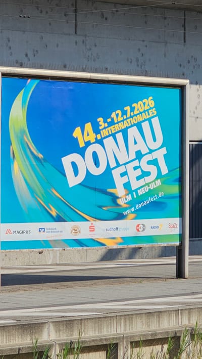 A large, rectangular billboard advertises the "DONAU FEST" in Ulm and Neu-Ulm. The billboard is predominantly blue and teal, with a dynamic, abstract design of yellow and green swirls on the left side. Prominently displayed in large white and yellow lettering are the words "14. DONAU FEST". Below this, "3.-12.7.2026 INTERNATIONALES" is written, followed by "ULM I NEU-ULM" and the website "www.donaufest.de".
At the bottom of the billboard, a row of sponsor logos is visible. These include "MAGIRUS", "Volksbank Ulm-Biberach eG", a circular logo with a lion, "Sparkasse Ulm", "sudhoff gruppe", and logos for "RADIO 7" and "SpaZz". The billboard is mounted on a metal frame and supported by poles, positioned outdoors in front of a concrete wall. The ground in front of the billboard is a paved walkway with horizontal lines. The scene appears to be a public outdoor space, likely in the city of Neu-Ulm, Germany, given the text on the advertisement. There are no people visible in the image. The environment is well-lit, suggesting daytime.