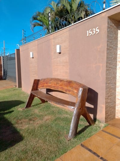 The image shows a rustic wooden bench placed on a lawn in front of a building with a brown stucco wall. The building has two square white lights mounted on the wall, and the address number "1535" is visible in white on the upper right portion of the wall. Palm trees and barbed wire are visible above the wall, suggesting a sunny outdoor environment. To the left of the bench, a metal gate is partially visible. The ground in front of the building is paved with tan square tiles. The scene appears to be an exterior of a residential or commercial property in São Gabriel do Oeste, Brazil.