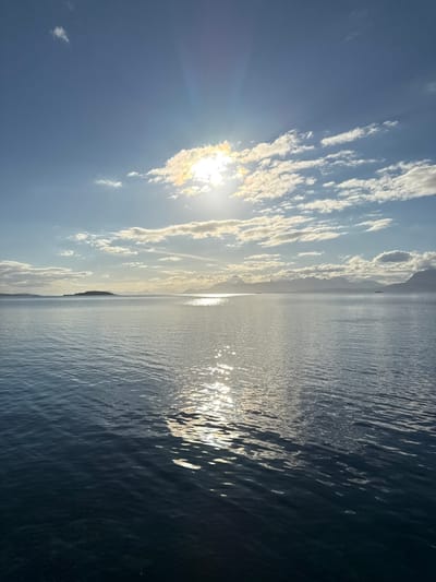 The image shows a wide expanse of calm, deep blue water, likely the sea, under a bright, partially clouded sky. The sun is high and shining, creating a shimmering reflection on the water's surface. In the distance, a low-lying island or peninsula is visible on the left, with a chain of mountains forming a silhouette against the horizon in the background. The overall scene evokes a sense of tranquility and natural beauty.
Based on the prominent mountain ranges and the characteristic coastline, this scene is likely located in or near Harstad, Norway. Harstad is situated in Troms og Finnmark county, known for its dramatic fjords, mountains, and coastal landscapes. The presence of islands and the vast body of water are consistent with the geography of the region. There are no people, specific objects, or actions depicted in the image; it is purely a landscape photograph.