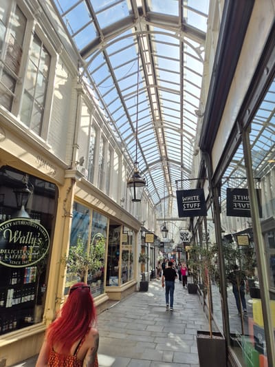 The image captures the interior of a shopping arcade in Cardiff, United Kingdom, characterized by a vaulted glass roof supported by a white metal framework. Shops line either side of the arcade. Signs for "White Stuff" and "Wally's Delicatessen & Kaffehaus" are visible. People are walking along the arcade's paved walkway. The architecture is a notable feature, with detailed building facades and decorative elements. The photograph has a perspective that emphasizes the length of the arcade. A woman with long red hair stands in the lower right corner.