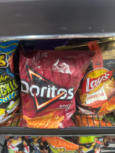 A close-up, eye-level shot shows a shelf stocked with various bags of chips in a retail setting. The lighting is artificial, and the shelf appears to be made of dark metal.

Dominating the center of the frame is a large, crinkled, red bag of Doritos, prominently displaying the brand's iconic logo. The text "SPICY NACHO" is visible in smaller lettering below the logo, and a graphic of flames and chip pieces adorns the bottom of the bag.

To the left of the Doritos bag, a portion of a bag of "Pork Rinds" is visible, featuring a bright yellow and blue design with text that includes "hot pickle puffs."

To the right of the Doritos bag is a bag of Lay's potato chips. The Lay's logo is clearly visible, along with the text "BARBECUE FLAVORED" and "MADE WITH REAL POTATOES." Behind the Lay's bag, another bag of chips, possibly also Lay's, with an orange and yellow design, is partially visible.

Below the main shelf, another shelf is visible, lined with more bags of chips, mostly in orange and yellow packaging, suggesting a variety of flavors. Some of these bags appear to be Cheetos.

The overall impression is that of a well-stocked snack aisle in a convenience store or supermarket. The specific location of Lake City, United States, is not discernible from the image itself.