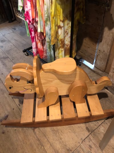 In a dimly lit store in Severn, Canada, a wooden rocking toy, shaped like a dinosaur with large ears and a smiling face, sits on a wooden floor. The toy is made of light-colored wood and has a saddle on its back. Behind the toy, a rack of brightly colored, tie-dyed t-shirts hangs, displaying vibrant hues of pink, orange, green, and yellow. The store appears to have rustic wooden walls and a wooden floor with visible grain and knots. The overall impression is one of a charming, handmade item displayed in a cozy, perhaps artisan, retail environment.