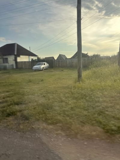 The image depicts a rural scene in Novonikolayevka, Russia, with a light-colored sedan parked on a grassy area next to a wooden fence. Several houses with dark roofs are visible behind the fence. A utility pole stands in the foreground, with power lines stretching across the sky, which is partly cloudy. The overall impression is that of a quiet, residential area on a bright day.