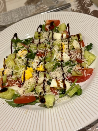 The image shows a close-up, overhead view of a white, ridged plate of salad. The salad consists of arugula, chopped tomatoes, cucumber slices, cubed mozzarella cheese, and hard-boiled egg wedges. It is topped with shredded Parmesan cheese and drizzled with a dark balsamic glaze. The salad is served on a table with a patterned placemat and a silver fork visible in the background. The setting appears to be indoors, possibly a restaurant or dining room, and the lighting suggests it could be evening. There are no people visible in the image. The location is not identifiable from the visual information provided in the image itself; therefore, it is not possible to determine if it is in Balbriggan, Ireland.
