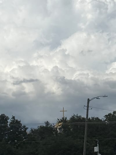 A golden cross is visible above a treeline under a cloudy sky. A wooden utility pole with a street light and wires is in the foreground. The image appears to be taken in North Port, United States of America.