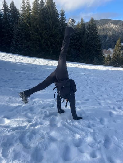 A solitary individual is depicted mid-cartwheel or similar acrobatic maneuver on a snow-covered slope in Saanen, Switzerland. The person wears dark winter clothing, including a jacket, pants, and hiking boots, and carries a black backpack. Their hands are planted in the snow, supporting their inverted body, with legs extended vertically upwards, one leg slightly higher than the other. The immediate ground is covered in white snow, displaying various footprints and subtle depressions. In the background, dense evergreen forests cover ascending slopes, with distant snow-capped mountains visible under a bright, partially cloudy blue sky.