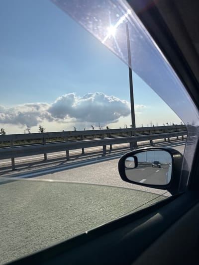 This image depicts a view from inside a vehicle traveling on a highway in Surakhany Raion, Azerbaijan. The perspective is from the passenger side window, looking out. The scene captured is primarily the sky, road, and a side-view mirror.

The sky is a clear, bright blue with several large, fluffy white clouds scattered across it, particularly on the left side of the frame. The sun is visible in the upper left portion of the image, casting a bright glare and lens flare onto the window. A metal guardrail runs parallel to the road, separating the highway lanes from the adjacent landscape. Beyond the guardrail, there are hints of vegetation and trees, suggesting an outdoor, possibly rural or semi-rural, setting.

The side-view mirror is prominently featured in the right half of the image. It reflects the road ahead, showing another vehicle traveling in the same direction. A smaller, convex mirror is attached to the main mirror, offering a wider field of view. The road itself appears to be paved and marked with white lane lines.

The overall impression is of a sunny day, likely mid-morning or early afternoon, given the brightness of the sun and the clear sky. The image evokes a sense of movement and travel. There is no visible text in the image. The scene is mundane and typical of a road trip or commute.